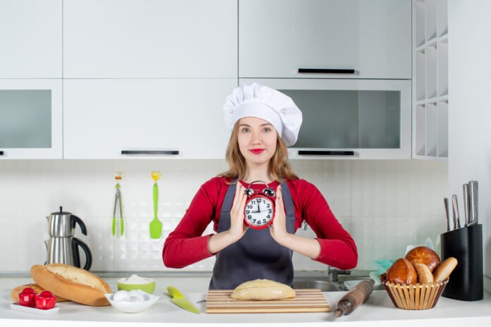 Chef segurando relógio enquanto prepara pão na cozinha para ilustrar dicas de economia de tempo no preparo de refeições.