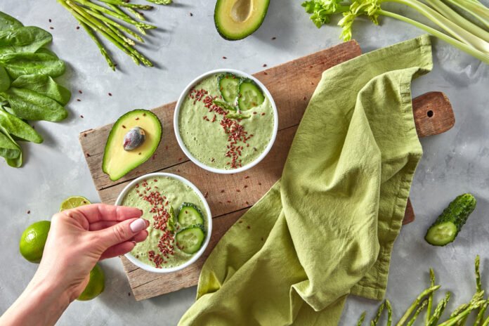 Tigela com sopa verde vegana de abacate e pepino, decorada com sementes de linhaça, acompanhada de vegetais frescos.