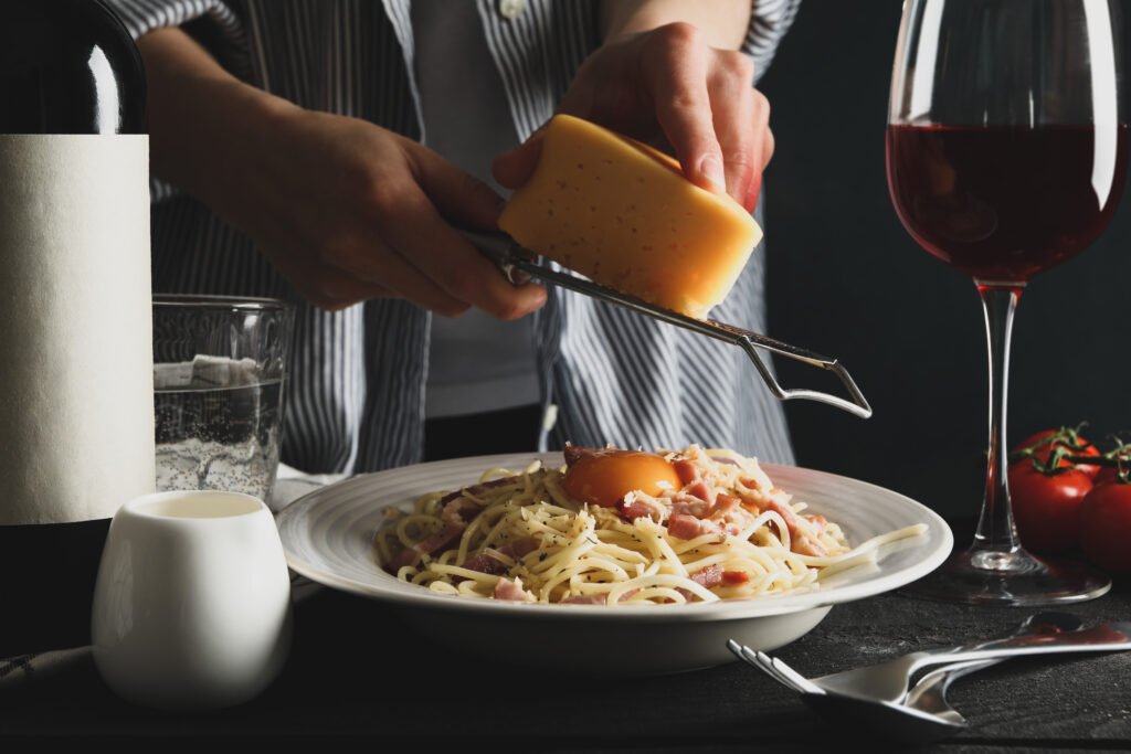 Pessoa ralando queijo sobre prato de espaguete carbonara com taça de vinho tinto.