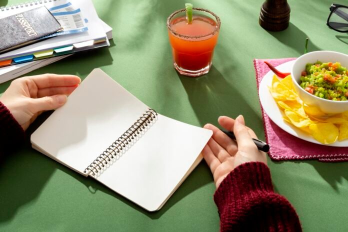 Mão segurando um caderno aberto ao lado de um prato de comida e uma bebida, simbolizando o planejamento de refeições semanais.