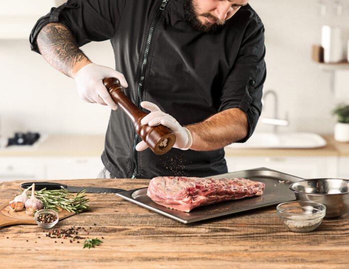 Chef temperando carne com pimenta e ervas frescas para preparo perfeito.