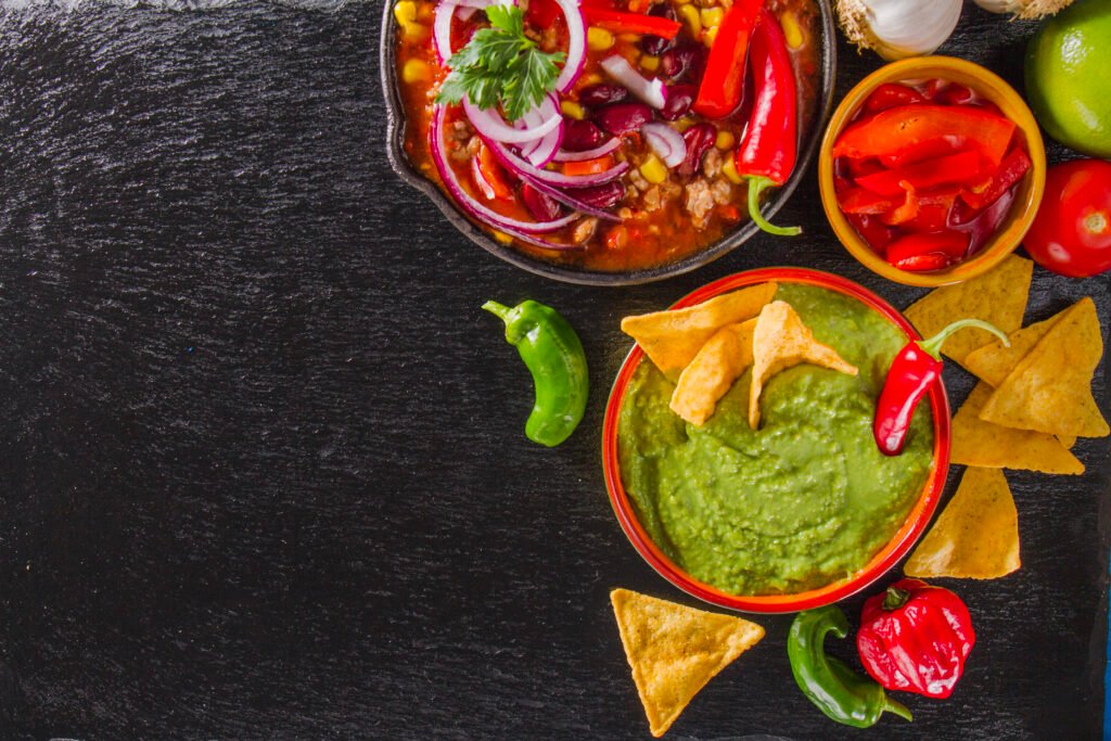 Pratos mexicanos com guacamole, pimenta e nachos sobre fundo preto.