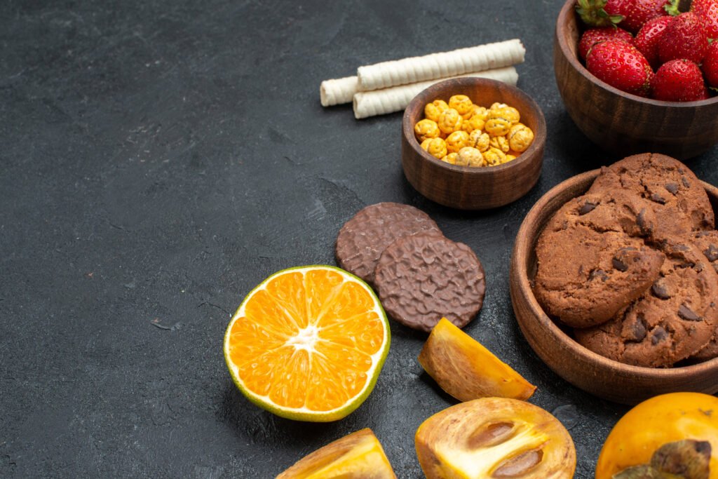 Doces saudáveis e frutas frescas como cookies integrais, tangerina, morangos e caqui em uma mesa com fundo escuro.