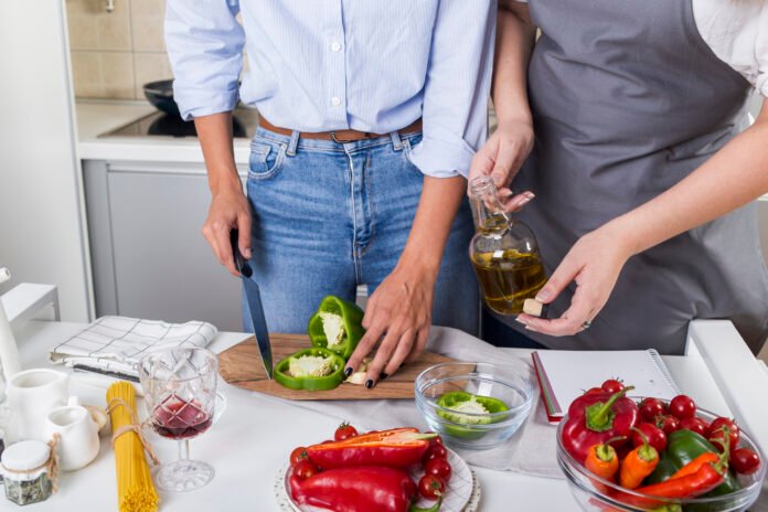 Duas pessoas preparando uma refeição, cortando pimentão verde e adicionando azeite em uma cozinha.