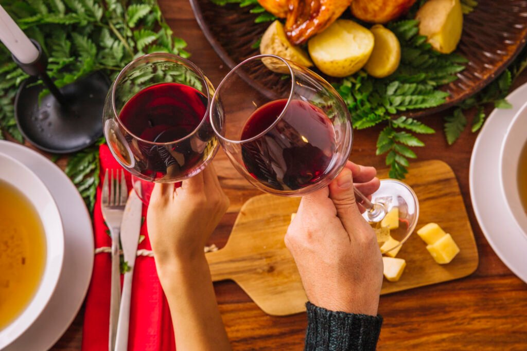 Duas taças de vinho tinto brindando em uma refeição elegante com queijo e pratos variados na mesa.