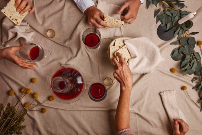 Pessoas compartilhando vinho e pão em um ambiente aconchegante, com elementos de decoração e guardanapos sobre a mesa.