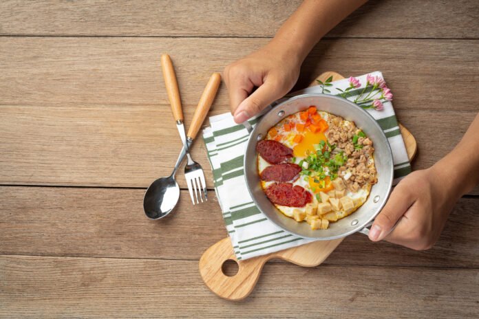 Prato de café da manhã com ovo, legumes e linguiça, servido em uma frigideira pequena sobre uma tábua de madeira.