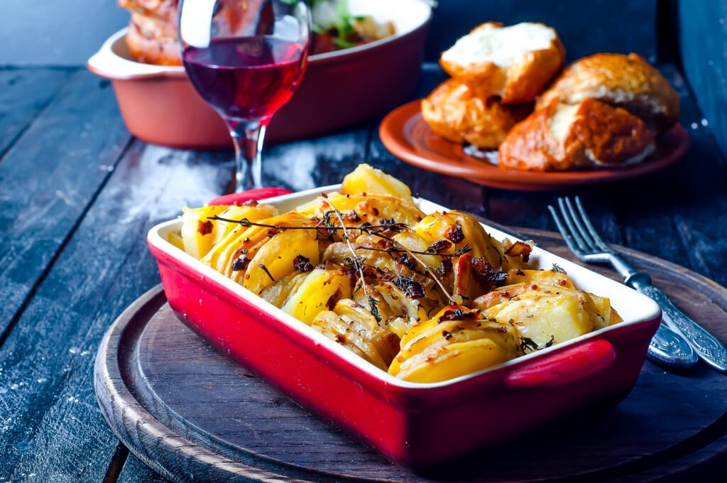 Prato de batatas assadas com ervas em uma travessa vermelha, acompanhado de uma taça de vinho tinto e pães ao fundo.