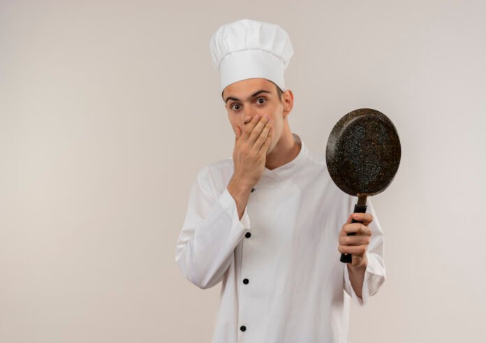 Chef surpreso segurando uma frigideira, vestido com uniforme branco de chef e chapéu.