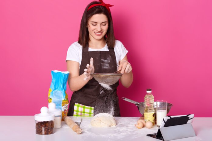 Jovem confeiteira preparando massa para bolo em bancada com ingredientes, sorrindo enquanto peneira a farinha.