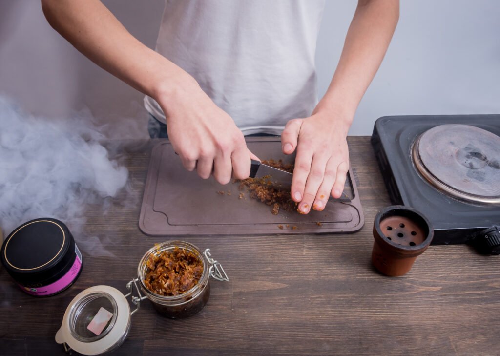 Pessoa picando ingredientes defumados em uma tábua de corte com jarra e fogão ao fundo.