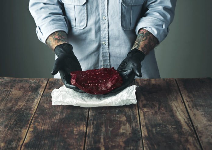 Chef segurando carne crua com luvas pretas, pronta para selagem e defumação.