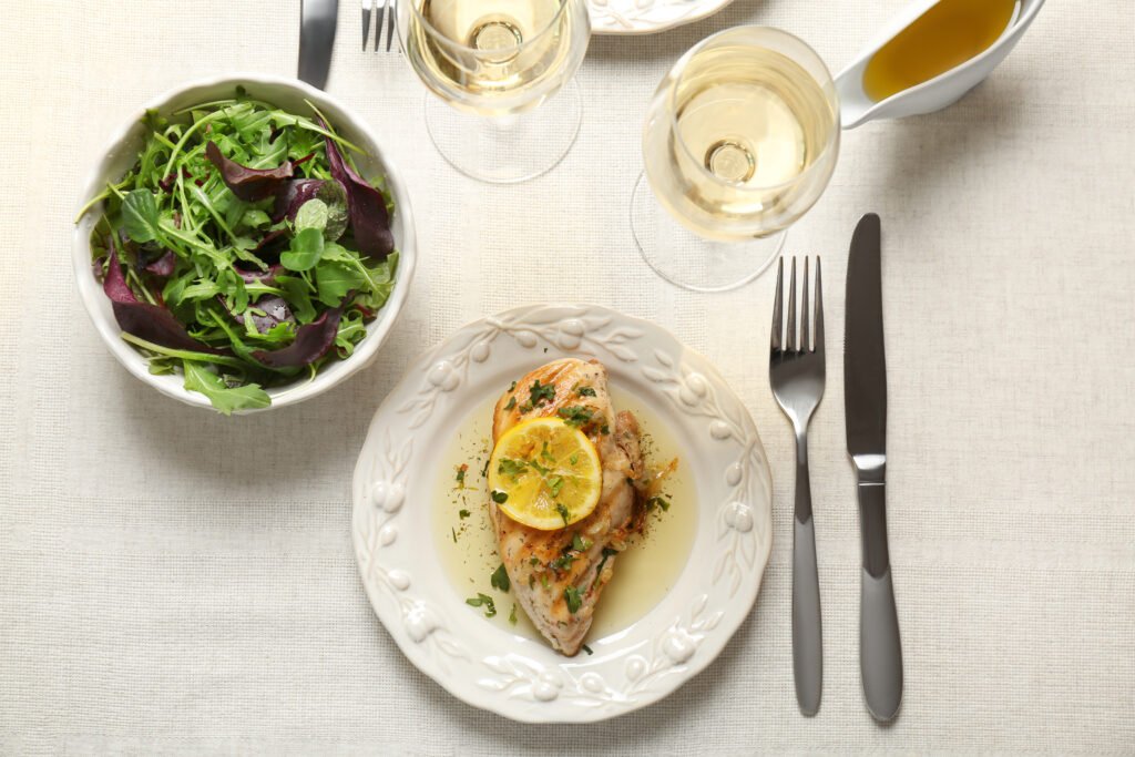 Prato de frango com molho de limão, acompanhado de salada e taças de vinho branco, em uma mesa elegante.