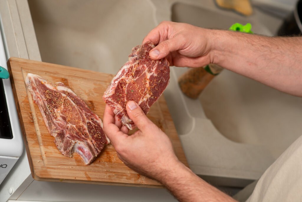 Pessoa temperando pedaços de carne em uma tábua de madeira, preparando para cozimento.