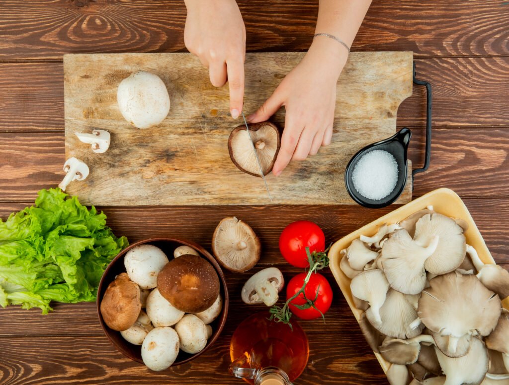 Pessoa cortando cogumelos frescos em uma tábua de madeira com ingredientes como alface, tomate e sal ao redor.