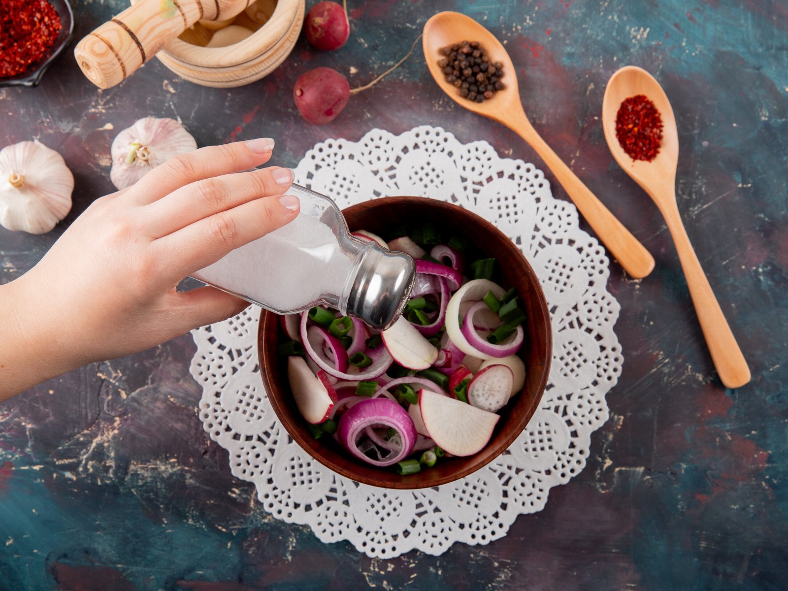 Mão adicionando sal a uma tigela com vegetais frescos, rodeada de temperos como alho e pimenta.