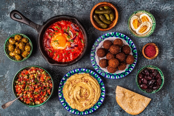 Uma mesa repleta de pratos tradicionais da cozinha árabe, incluindo falafel, homus, shakshuka, salada tabule, pães e azeitonas.