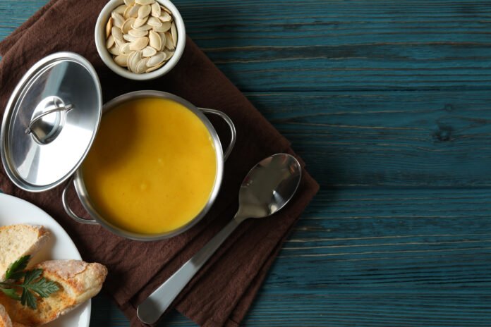 Tigela de caldo caseiro amarelo em uma mesa azul com colher e pão como acompanhamento.