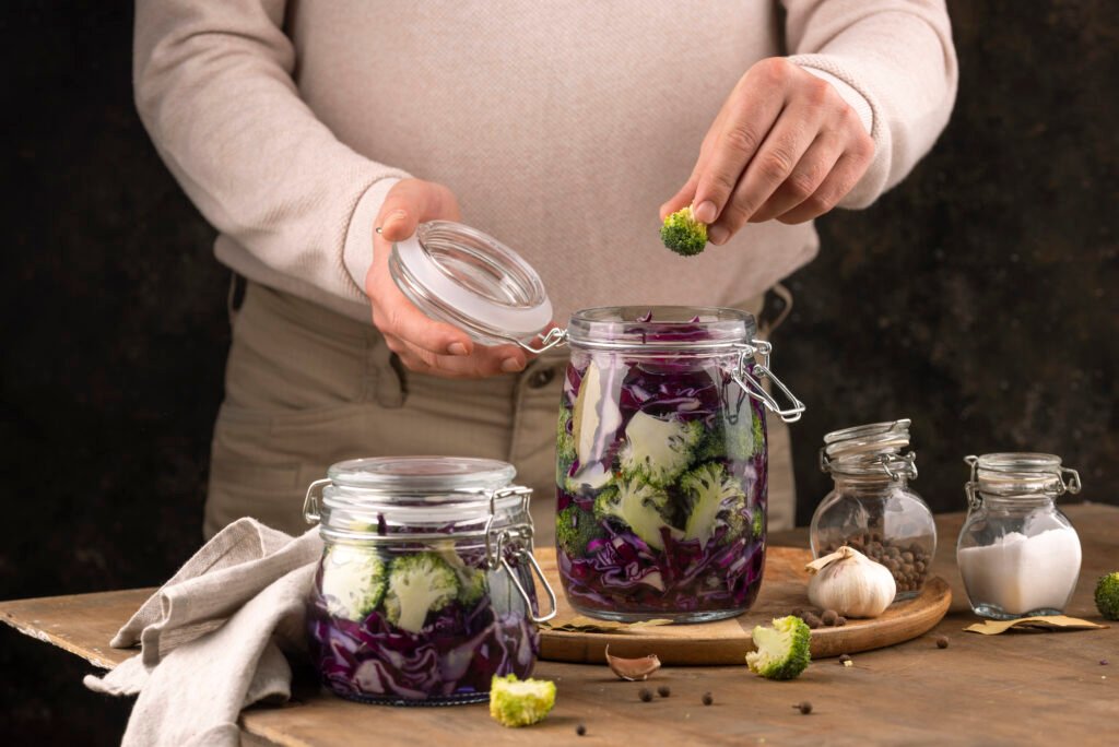 Pessoa colocando um pedaço de brócolis em um pote de vidro com vegetais fermentados, incluindo repolho roxo e couve-flor, em uma mesa de madeira com temperos ao redor.